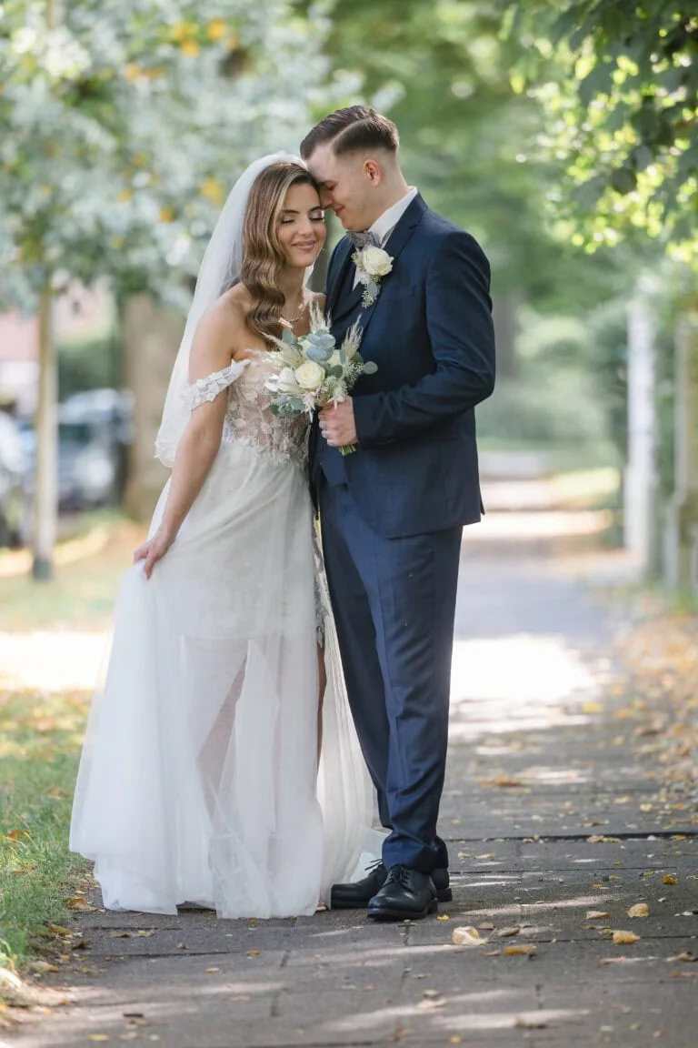 Ein Brautpaar steht auf einem Weg, umgeben von Grün. Die Braut trägt ein schulterfreies Kleid mit Schleier und hält den Rock des Kleides, der Bräutigam hält den Brautstrauß. Sie lehnen ihre Köpfe aneinander und genießen den Moment.