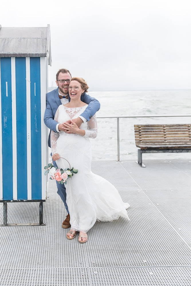 Brautpaar steht lachend an ein Badehäuschen gelehnt auf einem Steg, im Hintergrund sieht man die aufgewühlte Ostsee in Stein bei Laboe, fotografiert von Photography Team Hochzeitfotograf Kiel