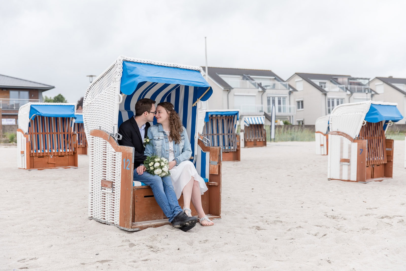 Brautpaar sitzt in einem weißen Strandkorb am Strand von Sierksdorf, lächelt sich verliebt an, Brautstrauß auf dem Schoß der Braut, weitere Strandkörbe im Hintergrund