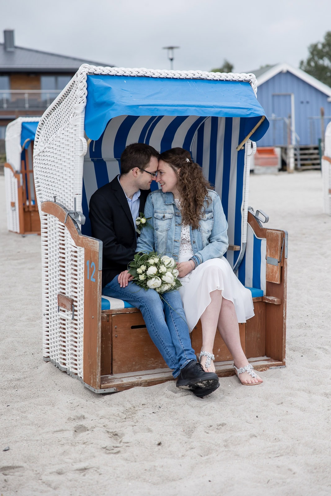 Brautpaar sitzt in einem weißen Strandkorb am Strand von Sierksdorf, legt die Stirn aneinander, Brautstrauß auf dem Schoß der Braut, weitere Strandkörbe im Hintergrund