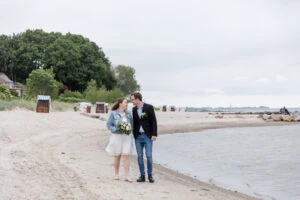 Brautpaar spaziert Hand in Hand am Strand in Sierksdorf, Braut trägt kurzes Kleid mit Jeansjacke und hält den Strauß, beide verliebt, Meer im Hintergrund – Hochzeit am Meer, Schleswig-Holstein